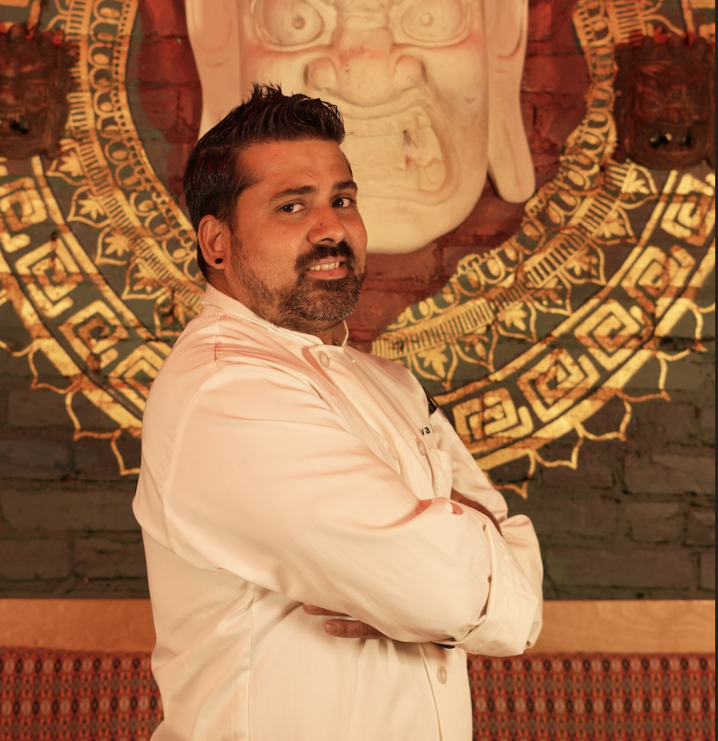 Chef in white uniform standing confidently in front of a decorative Nepalese mural